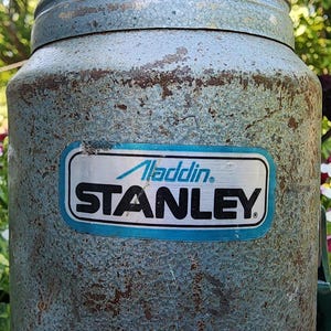May include: Vintage Aladdin Stanley thermos in a weathered, light blue finish. The thermos features a rectangular label with the "Aladdin" and "STANLEY" brand names in a blue and white design. The background includes blurred greenery and flowers.