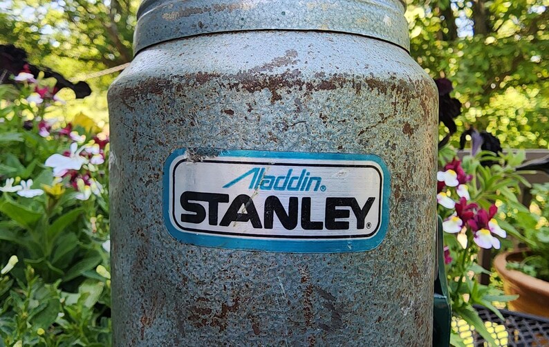 May include: Vintage Aladdin Stanley thermos in a weathered, light blue finish. The thermos features a rectangular label with the "Aladdin" and "STANLEY" brand names in a blue and white design. The background includes blurred greenery and flowers.