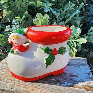 May include: A white ceramic Christmas boot planter with a red rim and a red and green elf figure on the side. The boot has a green holly leaf design with red berries.