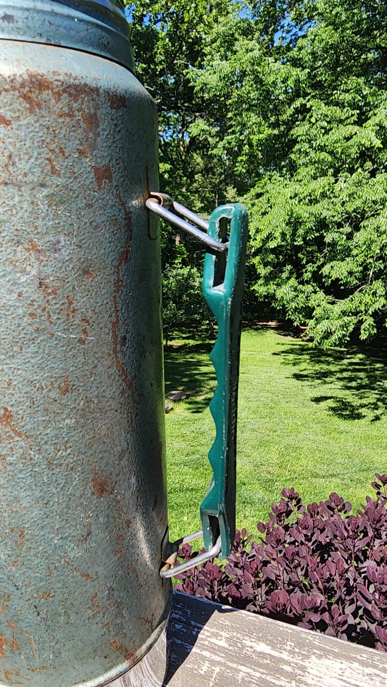 May include: Close-up of a vintage, weathered metal container with a green handle. The container has a textured, teal finish with rust spots. The handle is a dark green color and has a ridged design. The container is outdoors.