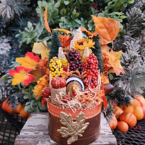May include: A small, round, brown container with an orange ribbon and a gold glitter leaf design. The container is filled with fall-themed decorations, including a miniature turkey, corn, pumpkins, and berries.