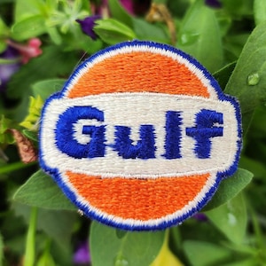 May include: A circular embroidered patch featuring the Gulf logo. The design consists of a bright orange circle bisected by a horizontal white band with a blue border. The word Gulf is stitched in bold blue letters across the white center.
