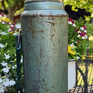May include: Vintage green thermos with a shiny silver lid and handle. The thermos has a textured surface and shows signs of wear. A classic outdoor item for keeping beverages hot or cold.