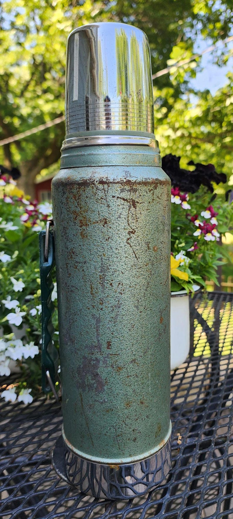 May include: Vintage green thermos with a shiny silver lid and handle. The thermos has a textured surface and shows signs of wear. A classic outdoor item for keeping beverages hot or cold.