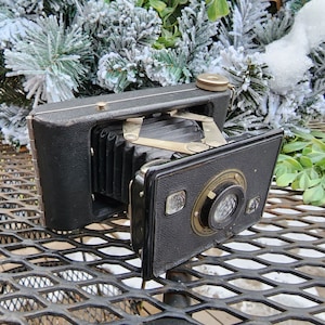 May include: A vintage black folding camera with a bellows and a lens. The camera is sitting on a metal table with a grid pattern.