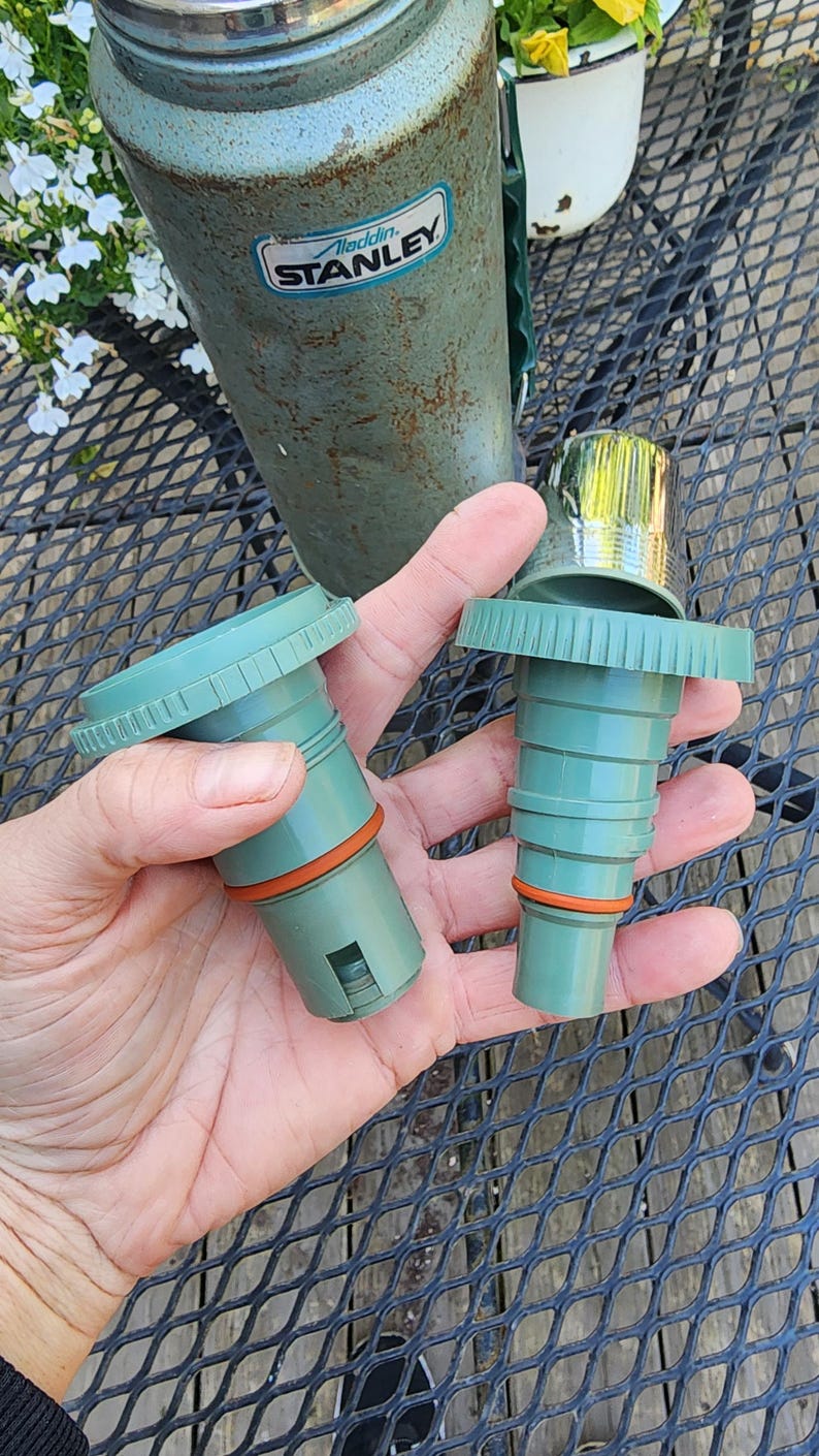 May include: Two green replacement stoppers for a vintage Aladdin Stanley thermos. The stoppers have an orange rubber seal and a ridged design. The thermos is visible in the background, with the Aladdin Stanley logo.
