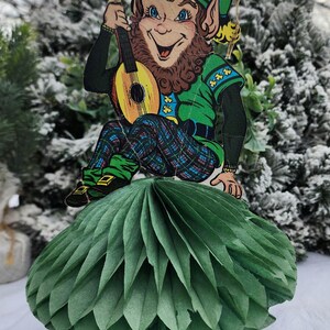 May include: A whimsical St. Patrick's Day decoration featuring a leprechaun with a guitar, seated atop a green paper honeycomb tree. The leprechaun is dressed in green and blue plaid, with a smiling face. The background is a snowy, blurred scene.