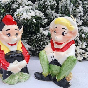 May include: Two ceramic figurines of elves. One elf is wearing a red jacket, black pants, and a red hat. The other elf is wearing a white shirt with a red collar, green pants, and a white hat. The figurines are on a snowy surface with a frosted evergreen background.