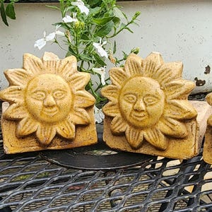 May include: Four decorative, sun-shaped garden stones. Each stone features a smiling sun face with petal-like rays, in a warm, earthy yellow-brown color. The stones are arranged on a black metal surface.