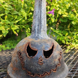 May include: A weathered, dark metal Halloween decoration with a jack-o'-lantern face. The sculpture has a tall, cylindrical neck and a bulbous base with cut-out eyes and a jagged mouth. The metal has a rusty, aged appearance.