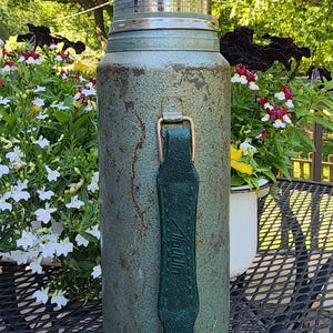 May include: Vintage green thermos with a shiny silver lid and a green leather handle. The thermos has a weathered appearance, suggesting age and use. A classic item for outdoor adventures or picnics.