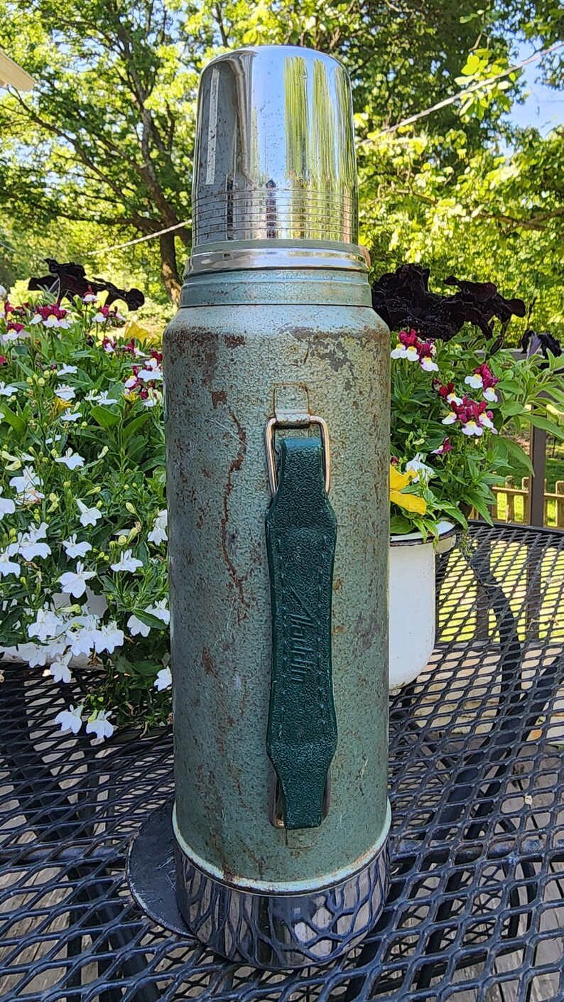 May include: Vintage green thermos with a shiny silver lid and a green leather handle. The thermos has a weathered appearance, suggesting age and use. A classic item for outdoor adventures or picnics.