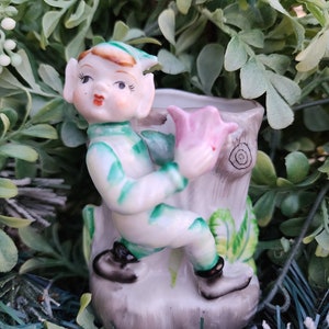 May include: A vintage ceramic figurine of an elf climbing a tree stump, holding a pink flower. The elf is wearing a green and white outfit with black shoes. The tree stump is gray with green leaves and a wood grain design. The figurine is surrounded by green foliage.