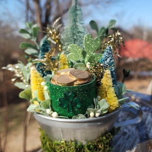 May include: A St. Patrick's Day decoration featuring a galvanized metal container filled with gold coins, a green glitter pot with a shamrock, and miniature trees. The arrangement is surrounded by greenery and moss.