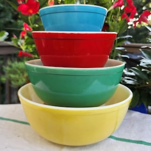 May include: A stack of four round mixing bowls in descending order of size from bottom to top. The bottom bowl is yellow, the second is green, the third is red, and the top bowl is blue. Each bowl has a smooth finish and a simple, rounded rim.