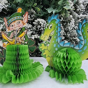 May include: Green St. Patrick's Day decorations featuring a leprechaun playing a flute and a cartoon dragon. The decorations include honeycomb paper shapes and are set against a snowy, green backdrop.
