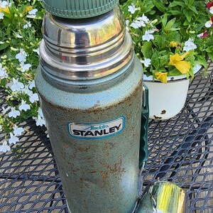 May include: Vintage Aladdin Stanley thermos in teal and silver. The thermos has a green handle and lid, with a matching cup. The Stanley logo is visible on the front. The thermos is sitting on a black metal table.