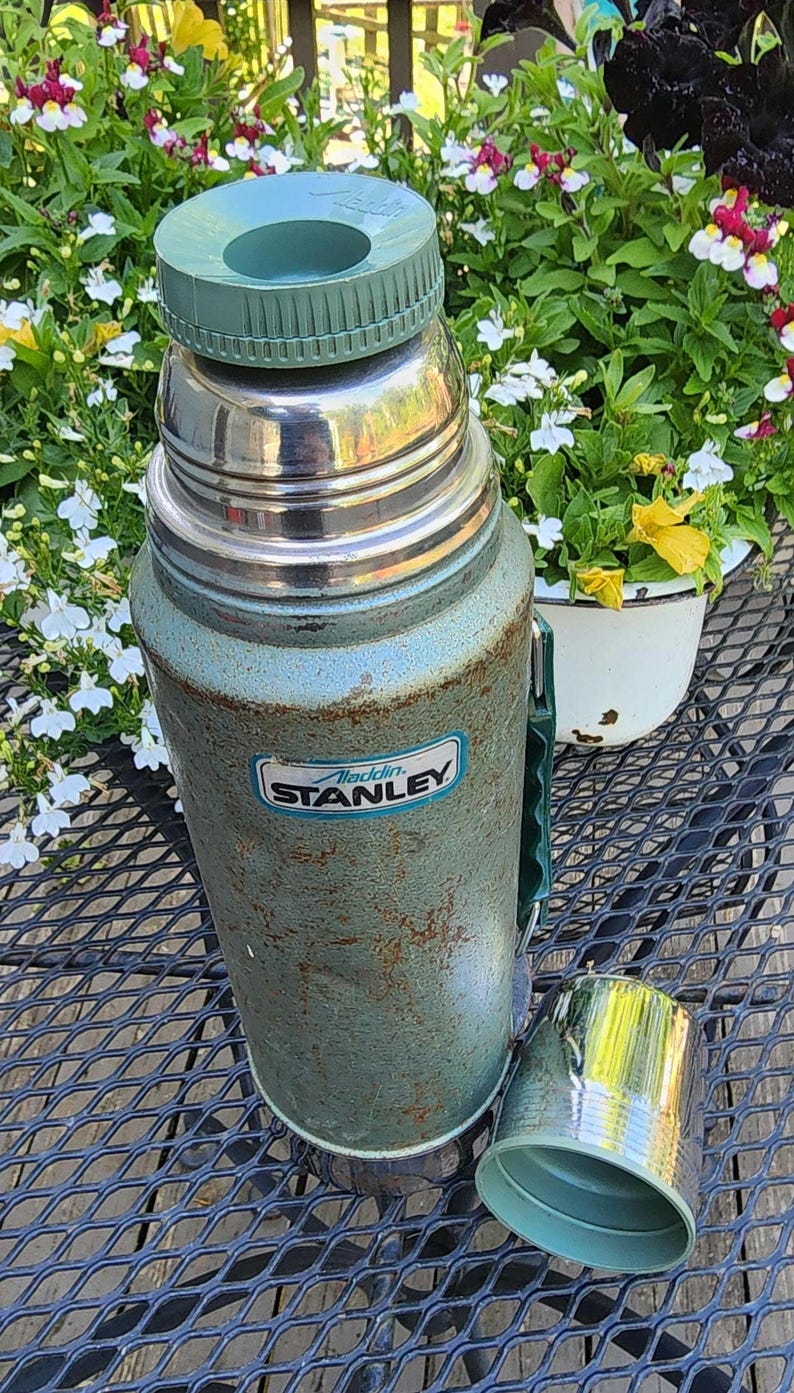 May include: Vintage Aladdin Stanley thermos in teal and silver. The thermos has a green handle and lid, with a matching cup. The Stanley logo is visible on the front. The thermos is sitting on a black metal table.