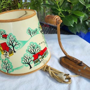 May include: A wall-mounted lamp featuring a conical cream-colored shade decorated with a painted landscape of houses, trees, and rolling hills in red, yellow, and teal. The lamp has a brown metal arm and a matching wall plate with a power cord.