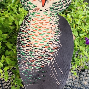 May include: A flat, die-cut owl wall decoration featuring a stylized design with orange, green, and black ink patterns. The owl is depicted with one large green eye open and the other closed in a wink, perched on a green branch with orange claws.