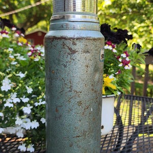 May include: Vintage teal Stanley thermos with a shiny silver lid and base. The thermos has a textured surface with signs of wear and tear. A classic item for outdoor adventures or retro decor.