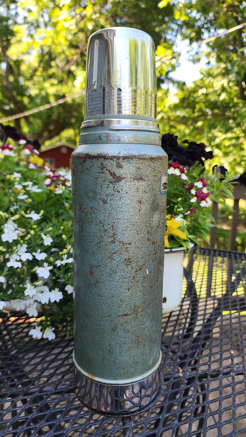 May include: Vintage teal Stanley thermos with a shiny silver lid and base. The thermos has a textured surface with signs of wear and tear. A classic item for outdoor adventures or retro decor.
