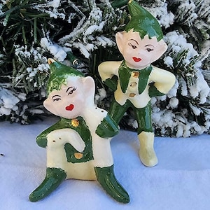 May include: Two vintage ceramic elf figurines. The elves have green hats and outfits with white accents. They have rosy cheeks, red lips, and are posed with their hands on their hips. The background features snowy evergreen branches, suggesting a holiday theme.