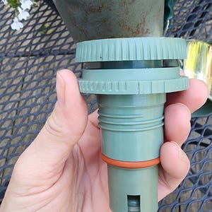 May include: A green and orange replacement part for a Stanley thermos. The part is cylindrical with a ridged top and an orange ring. The Stanley thermos is visible in the background.