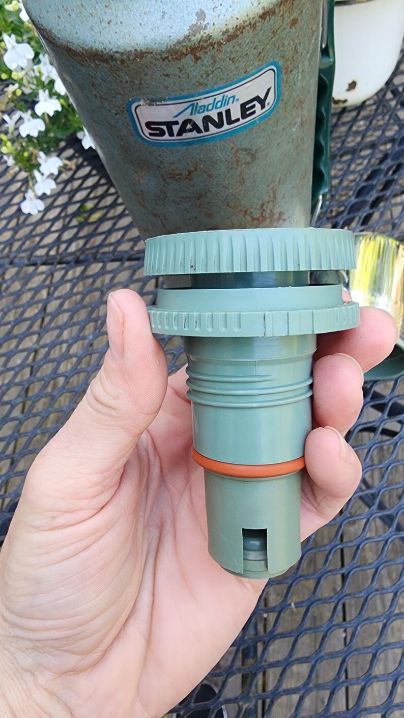 May include: A green and orange replacement part for a Stanley thermos. The part is cylindrical with a ridged top and an orange ring. The Stanley thermos is visible in the background.
