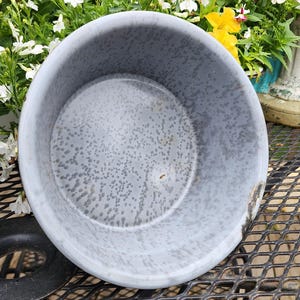 May include: A round, light gray enamelware bowl with a speckled pattern. The bowl has a slightly chipped rim and sits on a black metal surface. The background features colorful flowers.