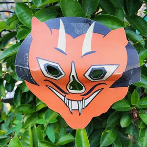 May include: A flat, orange paper mask featuring a stylized devil face with black accents, two white horns, pointed ears, and a wide grin showing sharp white teeth. The mask has cut-out eye holes and a triangular nose opening, with a smooth finish.