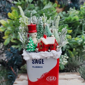 May include: A vintage red and white tin can with the words "Sage Rubbed" and "IGA" printed on it. The can is filled with a miniature winter scene featuring a snowman, a small house, and evergreen trees covered in white faux snow.