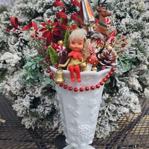 May include: A white milk glass vase filled with a festive Christmas arrangement. The arrangement includes red accents, an elf figurine, a reindeer, and various ornaments. The vase is decorated with a beaded garland and sits on a metal table.