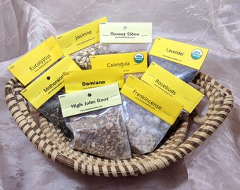 May include: A woven basket filled with various dried herbs and botanicals, including lavender, rosebuds, and frankincense. Each herb is labeled with a yellow tag. The basket is set against a soft, light-colored fabric background.
