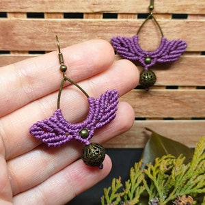 Könnte beinhalten: Lilafarbene Makramee-Ohrringe mit einem bronzefarbenen Metallperlen-Anhänger. Die Ohrringe sind handgefertigt und haben ein zartes, filigranes Design.