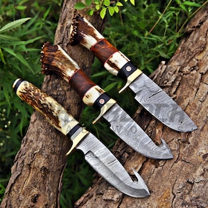 May include: Three fixed-blade knives with Damascus steel blades featuring a wavy pattern. Each knife has a handle made of textured brown antler and smooth dark wood, accented with a brass bolster and a small circular inlay on the handle.