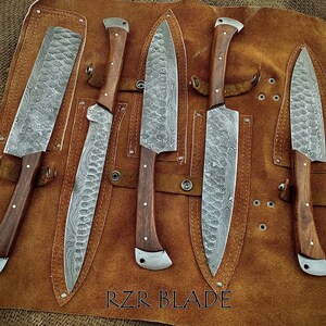 May include: Five Damascus steel kitchen knives with wooden handles are displayed in a brown leather roll. The knives are arranged in a row, with the blades facing up. The roll has a label that reads "RZR BLADE".