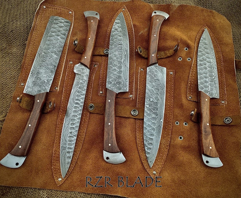 May include: Five Damascus steel kitchen knives with wooden handles are displayed in a brown leather roll. The knives are arranged in a row, with the blades facing up. The roll has a label that reads "RZR BLADE".