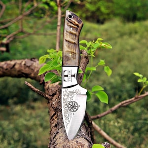 May include: A fixed-blade knife with a textured brown handle and a silver metal blade. The blade features an engraved compass design, topographic lines, and the text TYLER with a T-shaped logo. The handle includes a small hole at the top.
