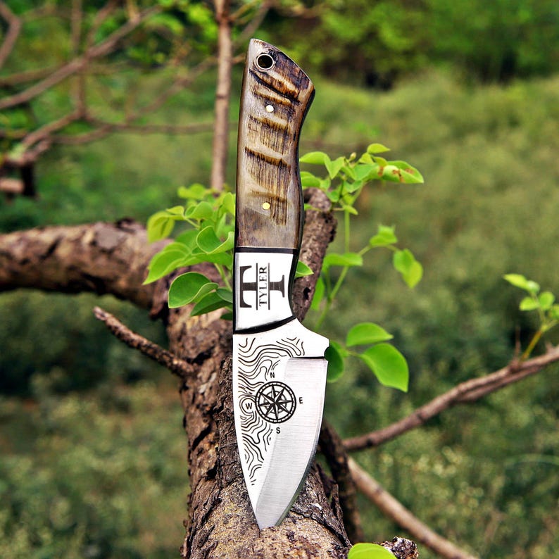 May include: A fixed-blade knife with a textured brown handle and a silver metal blade. The blade features an engraved compass design, topographic lines, and the text TYLER with a T-shaped logo. The handle includes a small hole at the top.