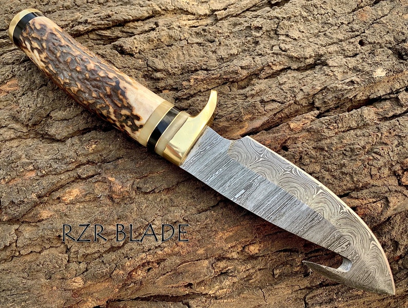 May include: A fixed-blade knife with a Damascus steel blade featuring a gut hook. The handle is crafted from textured brown stag horn with a brass guard and black spacers. The text RZR BLADE is visible on the left side of the blade surface.