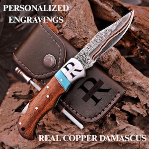 May include: A folding pocket knife with a patterned copper Damascus steel blade, a wooden handle, and a turquoise bolster. The bolster is engraved with the letters "RK" and the name "RALPH". It includes a brown leather sheath with "R" and "RALPH".