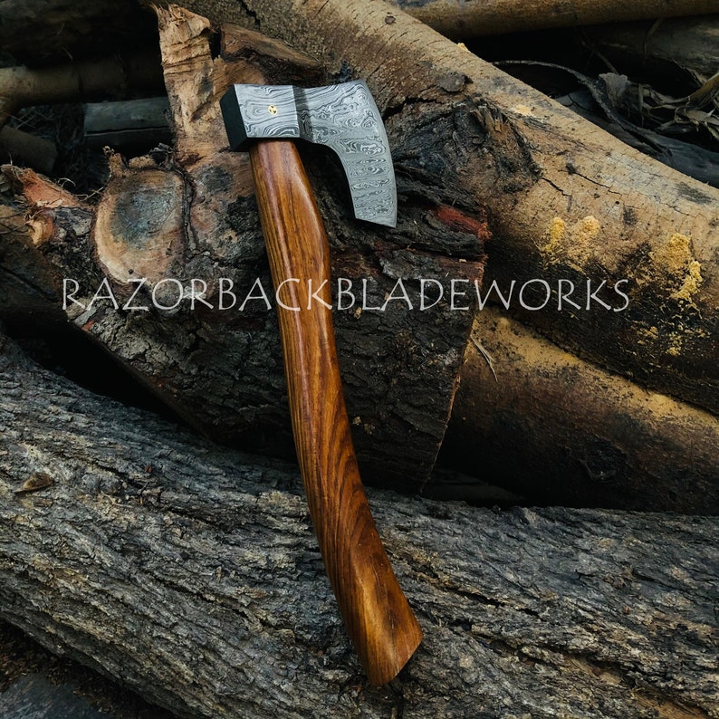 May include: A hand-forged Damascus steel axe with a wooden handle. The axe head has a unique pattern and is polished to a high shine. The handle is made of dark wood and has a smooth finish. The axe is resting on a pile of logs.