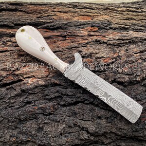 Peut inclure: Un couteau avec un manche en os blanc et une lame en acier damas. Le couteau est pos&eacute; sur un morceau de bois.