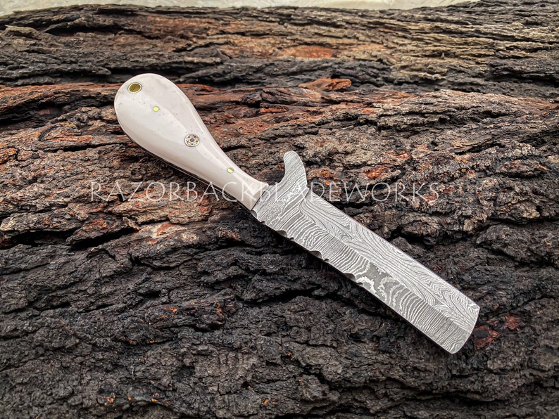 Peut inclure: Un couteau avec un manche en os blanc et une lame en acier damas. Le couteau est pos&eacute; sur un morceau de bois.