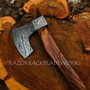 May include: A hand-forged axe with a Damascus steel blade and a wooden handle. The axe is resting on a wooden surface. The blade has a unique pattern and is very sharp. The handle is made of dark wood and is very sturdy. The axe is a beautiful piece of craftsmanship and would be a great addition to any collection.