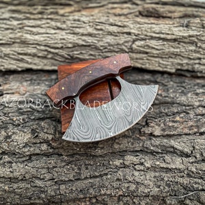 May include: A handcrafted Damascus steel alaskan ulu knife with a dark brown wooden handle. The blade features a unique pattern and is displayed on a wooden block.  The text 'RAZORBACKBLADEWORKS' is subtly visible. This high-quality, hand-forged knife is perfect for intricate carving tasks. The blade's curved shape and sharp edge are clearly visible, showcasing its functionality and craftsmanship.