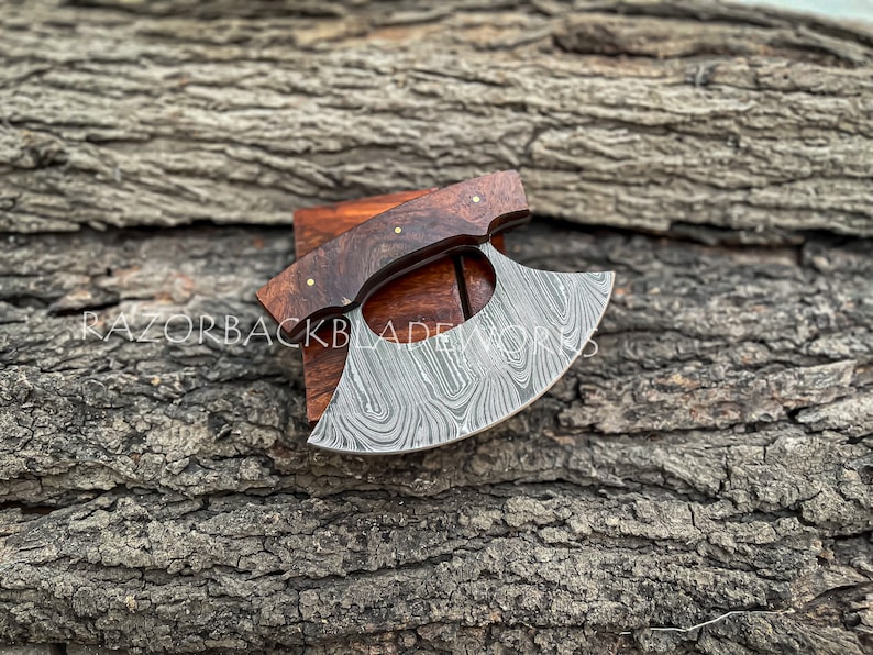 May include: A handcrafted Damascus steel alaskan ulu knife with a dark brown wooden handle. The blade features a unique pattern and is displayed on a wooden block.  The text 'RAZORBACKBLADEWORKS' is subtly visible. This high-quality, hand-forged knife is perfect for intricate carving tasks. The blade's curved shape and sharp edge are clearly visible, showcasing its functionality and craftsmanship.