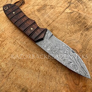 May include: A fixed-blade knife featuring a patterned Damascus steel blade and a dark brown wooden handle with horizontal grooves and two brass pins. The text RAZORBACK BLADE WORKS is visible on the blade and in the bottom left corner logo.