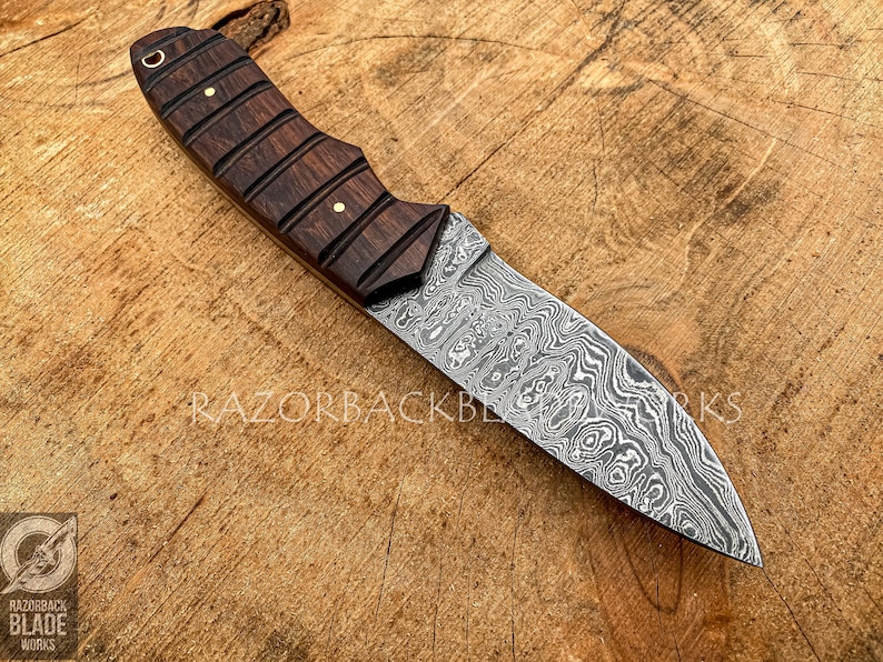 May include: A fixed-blade knife featuring a patterned Damascus steel blade and a dark brown wooden handle with horizontal grooves and two brass pins. The text RAZORBACK BLADE WORKS is visible on the blade and in the bottom left corner logo.